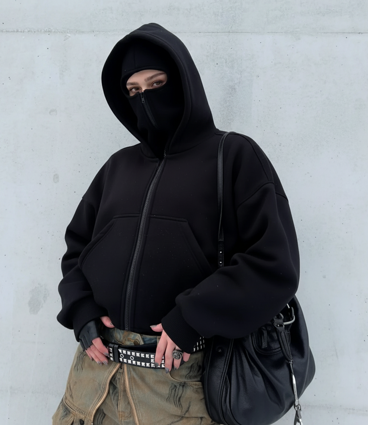 Person wearing a black hoodie and mask against a light gray wall.